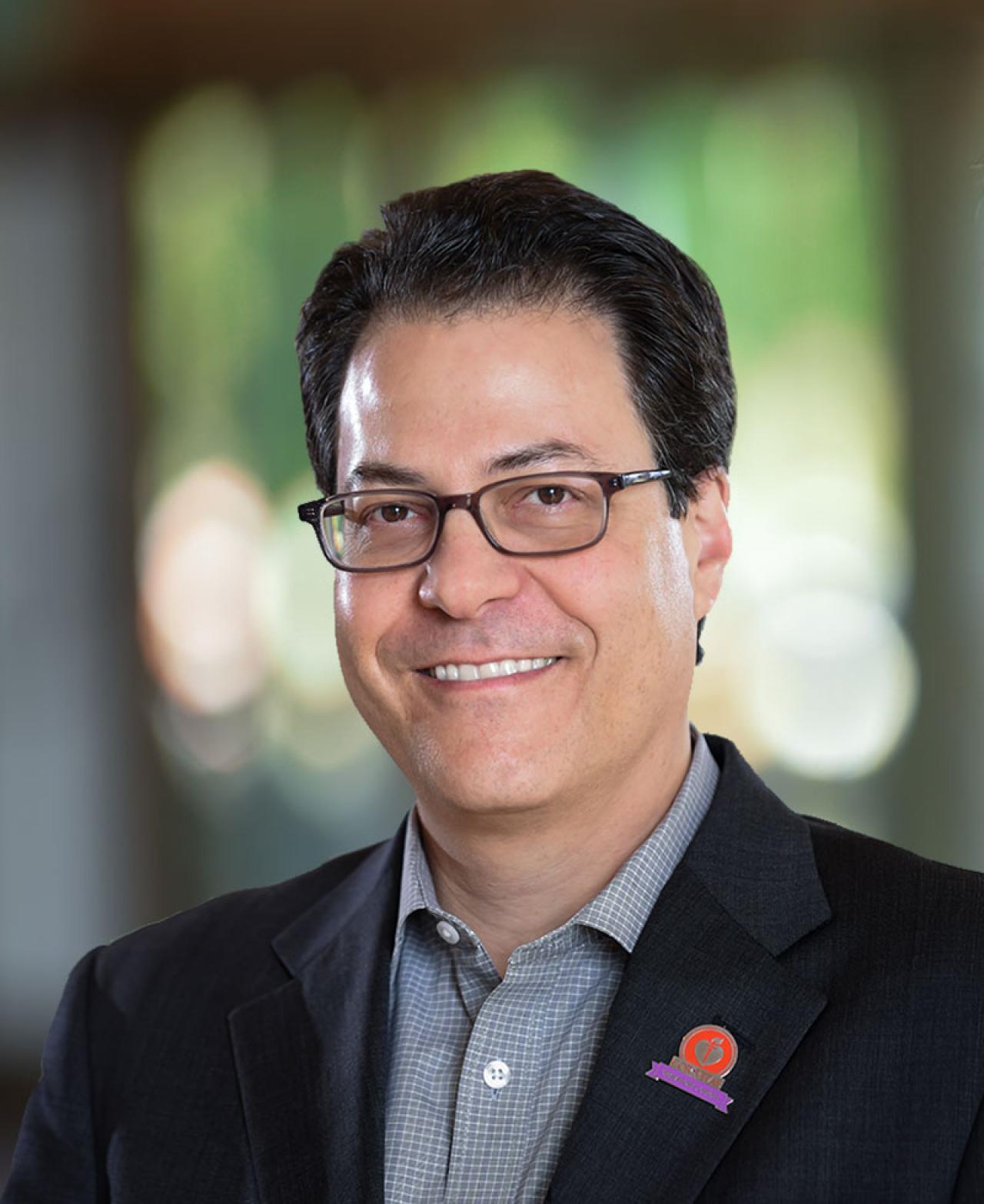 Photo of Mitchell Elkind, who wears a black suit jacket with a red pin on the lapel, with a grey shirt.