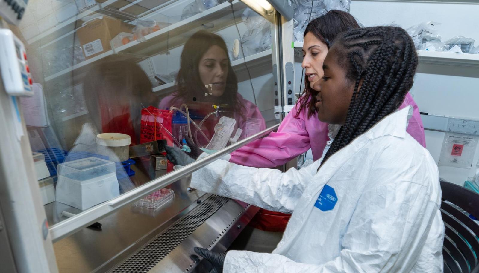 A woman in a pink lab gown helps a girl in a white lab coat use a pipette to transfer genetic material. 