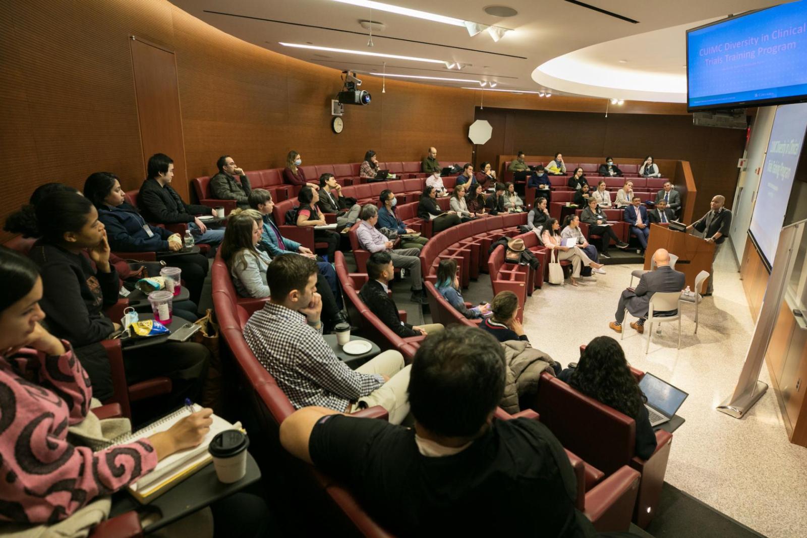 people in an auditorium watching and listening to a speaker at the front of the room