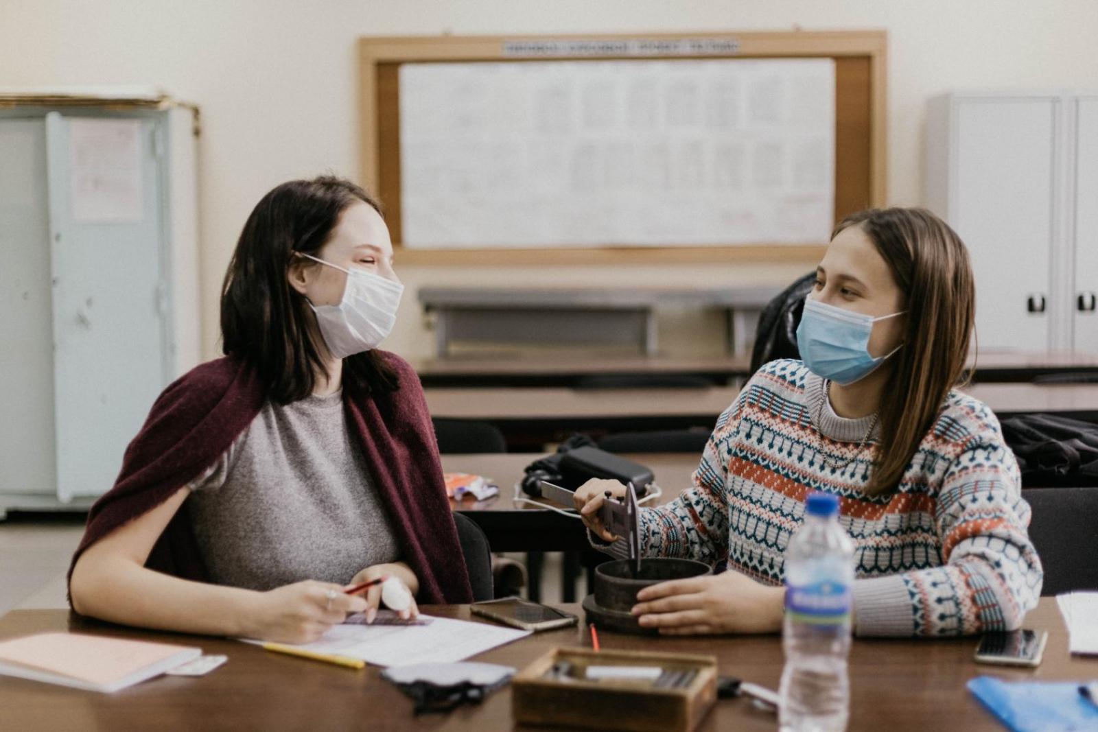 Two teenagers wearing face masks chat in a classroom