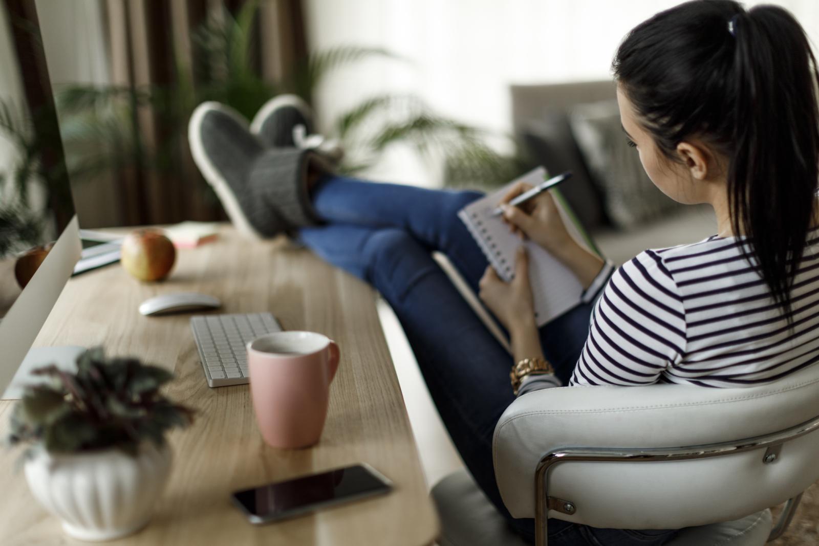 Young woman sitting at desk with feet up making list in notebook