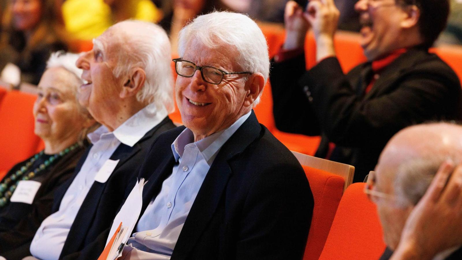 People sit in orange seats in an auditorium and laugh as they listen to a person speaking..