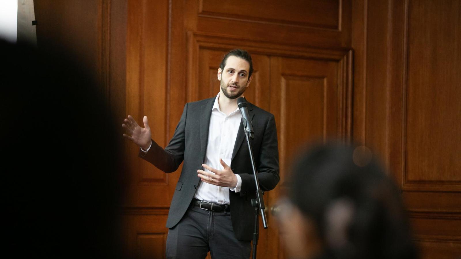 A man in a black suit stands behind a microphone and is speaking and gesturing towards a screen behind him