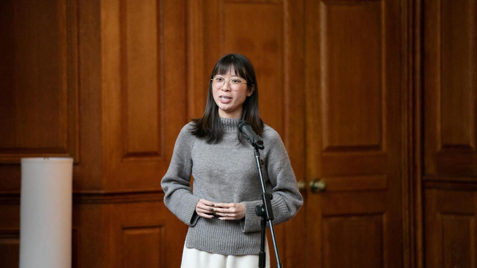 A woman in a grey sweater stands behind a microphone and is speaking