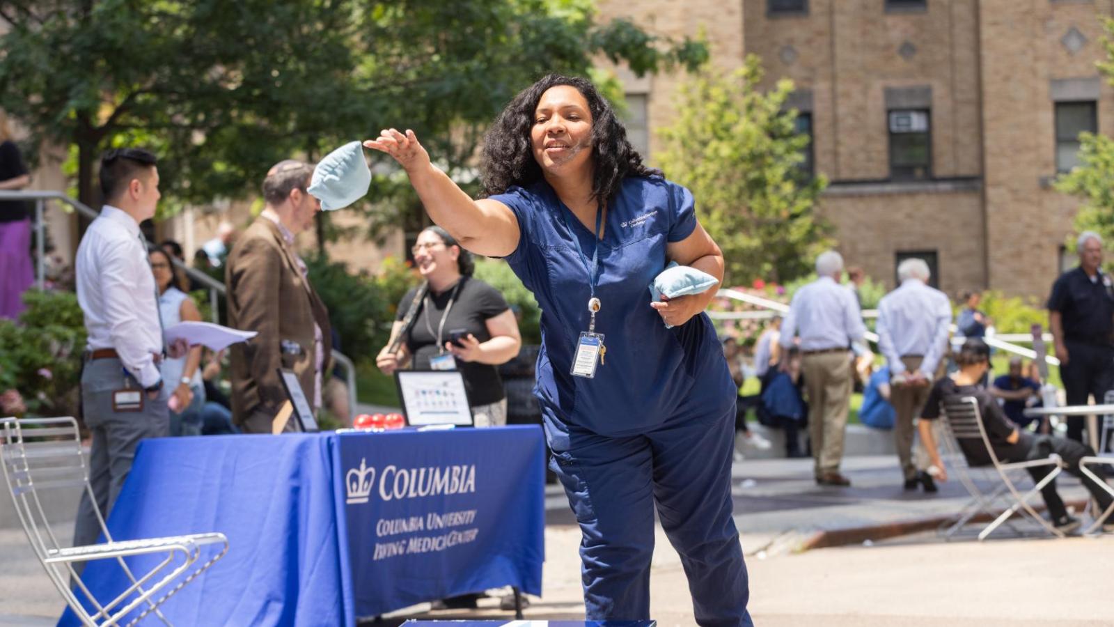A woman in blue scrubs tosses a bean bag