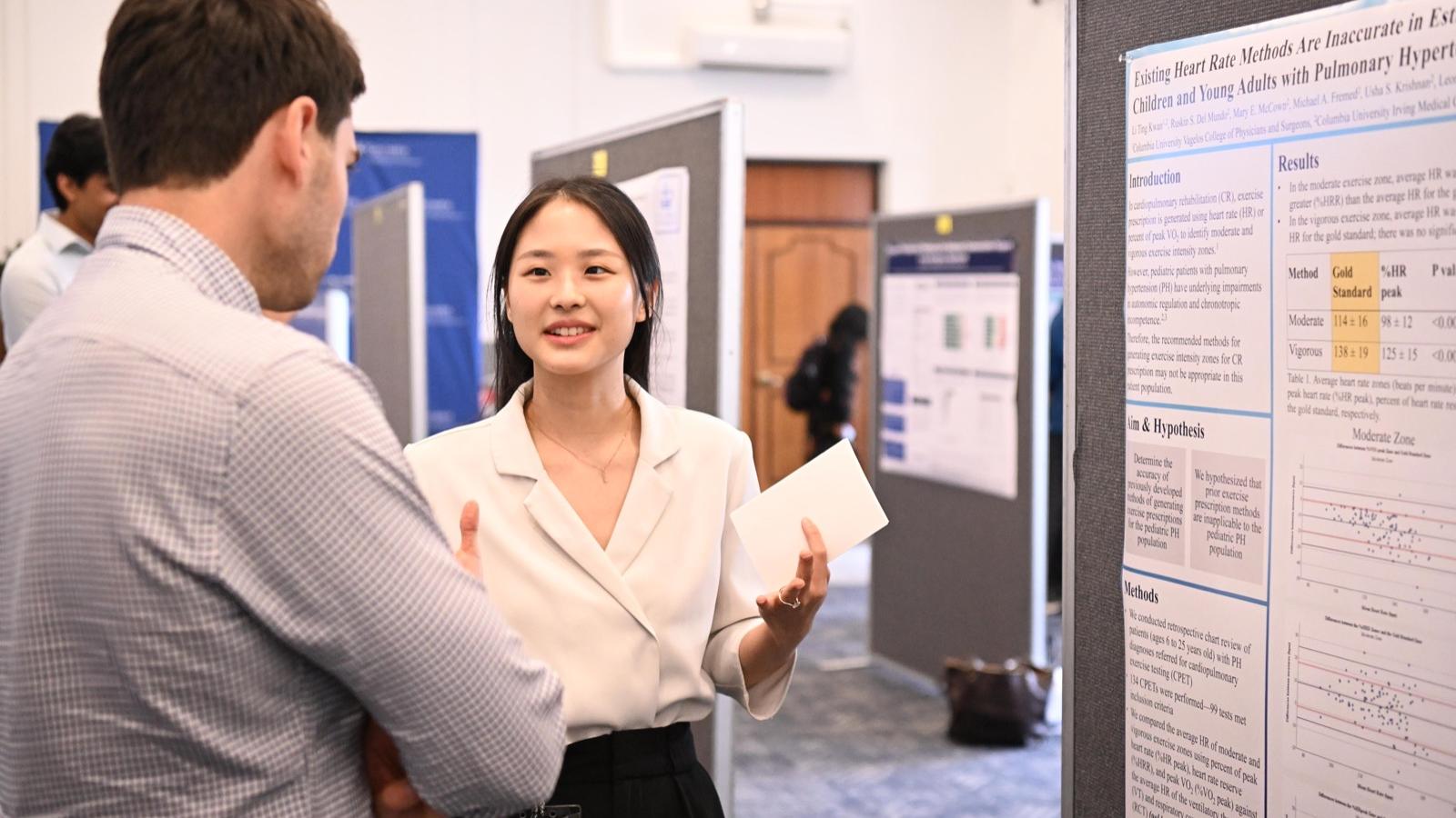 a student standing next to her research poster explains her findings to a judge
