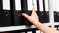 A bookshelf full of various binders with a person's arm pulling one of the binders off the shelf.