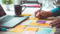 A person writing on stickies on a desk with a laptop