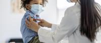Boy Receiving a Vaccination During a Pandemic. A sweet little mixed race boy sits up on an examination table as he receives an immunization. He is looking up and smiling as the doctor places the bandage on his arm.