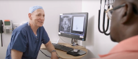 doctor in scrubs sitting in front of medical images on a computer and speaking to a patient