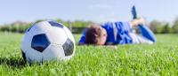 Male soccer (football) player in blue lying face down, injured on the field (pitch) on a clear day. Focus on traditional black and white ball.
