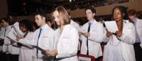 Students in white coats stand in an auditorium and read from a booklet.