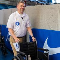 A man wearing a white polo shirt and blue button with the accessibility icon pushes a wheelchair
