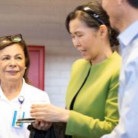 An older woman in a white polo shirt with reading glasses on her head smiles as a two people walk by her