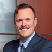 Dennis Scott-Torbet sits smiling while wearing a navy blue suit, with a light blue shirt and blue patterned tie